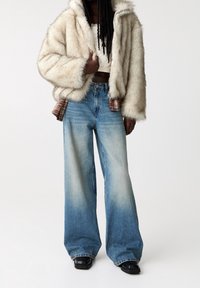 Veste en fausse fourrure ivoire avec capuche, associée à un jean flare bleu clair, présentant un léger dégradé et un détail écossais à la taille. Bottes noires.