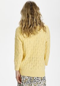 Gelber Strickpullover mit Rautenmuster, lockerer Passform, Dreiviertelärmel und geripptem Saum. Über einem Blumenkleid getragen.