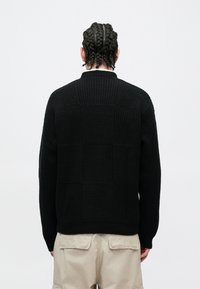 Pulover negru tricotat, cu model texturat în blocuri, guler și tiv reiat. Purtați cu pantaloni cargo bej, vizualizat din spate.