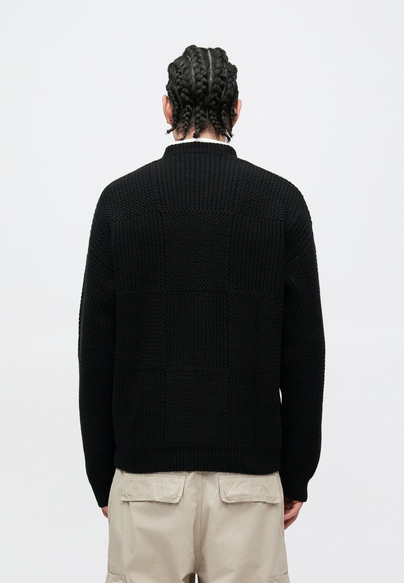 Sort strikket sweater med tekstureret blokkoste mønster, ribbet krave og kant. Bærer beige cargobukser, set bagfra.