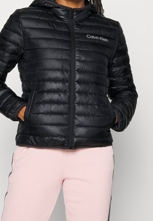 Zwarte doorgestikte Calvin Klein jas met rits aan de voorkant en capuchon, gedragen met lichtroze broek, handen in de jaspakken.