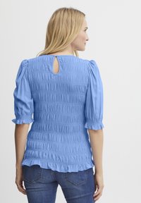 Lyseblå smock bluse med puffede ærmer, nøglehul tilbage og flæset kant. Materialet har et blødt, tekstureret udseende.