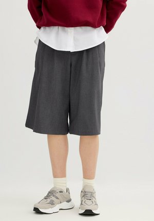 Persona con pantalones cortos de color gris oscuro hasta la rodilla, camisa blanca, suéter rojo, calcetines crema y zapatillas deportivas gris claro, de pie contra un fondo liso.