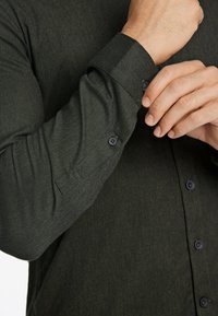 Camisa de botonadura completa de color verde oscuro con una tela texturizada, que presenta un puño de un solo botón y botones oscuros en contraste. Diseño ajustado y estrecho.