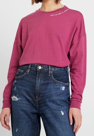 Person trägt ein langärmeliges magentarotes Shirt mit dem Text "LIFE'S TOO SHORT TO WASTE" nahe am Kragen und ausgewaschene blaue Jeans mit hoher Taille.