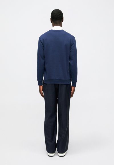 Άνδρας που στέκεται γυρισμένος, φορώντας σκούρο μπλε φούτερ, ριγέ σκούρο παντελόνι, με τον λευκό γιακά του πουκαμίσιού ορατό και λευκά αθλητικά παπούτσια, σε απλή φόντο.