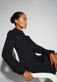 Zwarte op maat gemaakte jas met een kraag, knoopsluiting en zijzakken, gecombineerd met een bijpassende rok. Gladde textuur en gestructureerd ontwerp.