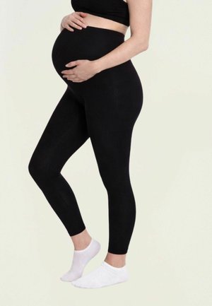 Personne enceinte portant des leggings de maternité noirs et des chaussettes blanches à la cheville, tenant son ventre avec les deux mains.