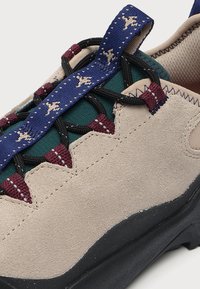 Chaussure de randonnée en daim beige avec semelle noire, œillets en tissu multicolores, lacets noirs et languette bleu avec petits motifs animaux beiges.
