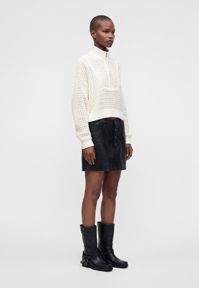 Femme portant un pull en maille blanc à demi-fermeture éclair, une mini-jupe en cuir noire et des bottes noires mi-mollet avec des détails de boucle, debout de profil.