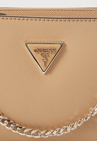 Beige strukturert veske med en fremtredende tredimensjonal logo som har "GUESS USA" i sentrum, aksentuert av en sølvkjede.