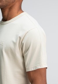 Close-up van een persoon die een lichtbeige katoenen T-shirt met korte mouwen draagt, op een effen lichtgrijze achtergrond.