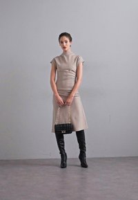 Beige farbendes, tailliertes Kleid mit kurzen Ärmeln, kombiniert mit einer schwarzen, strukturierten Handtasche und eleganten schwarzen Overknees. Glattes, minimalistisches Design.