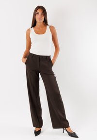 Pantaloni sartoriali marrone scuro con design a gamba dritta, tasche frontali e chiusura con un singolo bottone, abbinati a una canotta bianca.