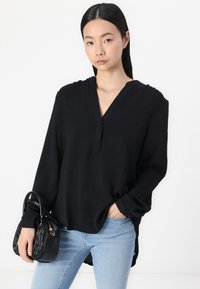 Ženska v ohlapni črni bluzici dolgimi rokavi in svetlo modrih kavbojkah, ki z eno roko drži majhno črno torbico z teksturo in ima drugo roko v žepu.