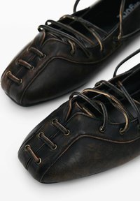 Black leather shoes featuring a rounded toe, prominent stitching, and lace-up design with gold-tone eyelets. Smooth texture and brown accents.