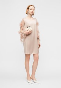 Φόρεμα κατσαρό χρώματος nude με διαφανείς, βολάν μανίκια. Το μοντέλο κρατάει μια κρέμ τσάντα φάκελο και φοράει λευκές γόβες με μυτερή μύτη. Απλός, κομψός σχεδιασμός.