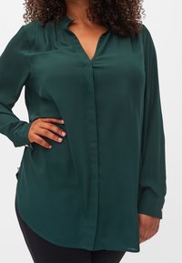 Blusa verde a maniche lunghe con scollo a V, texture liscia, orlo curvo e elementi decorativi ai polsini. Modello con una mano sulla vita.