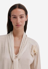 Beige blouse met textuur en zachte drapering, voorzien van een knoopsluiting bij de hals en een bloemenkantaccent op de schouder. Lichtgewicht stof.