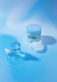 Klar glasflaske med Torriden-serum og pipette, omgivet af en blå krukke med Torriden Multi Pad og teal bomuldsprodukter på en blå baggrund.