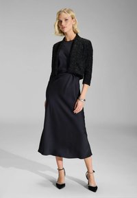 Vestido de noche de satén negro con una chaqueta corta y texturizada en negro. Presenta una silueta suave y fluida, acompañada de tacones negros de punta.