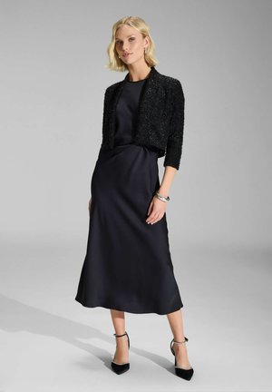 Robe de soirée en satin noir avec une veste tronquée texturée noire. Présente une silhouette lisse et fluide ainsi que des talons noirs à bout pointu.