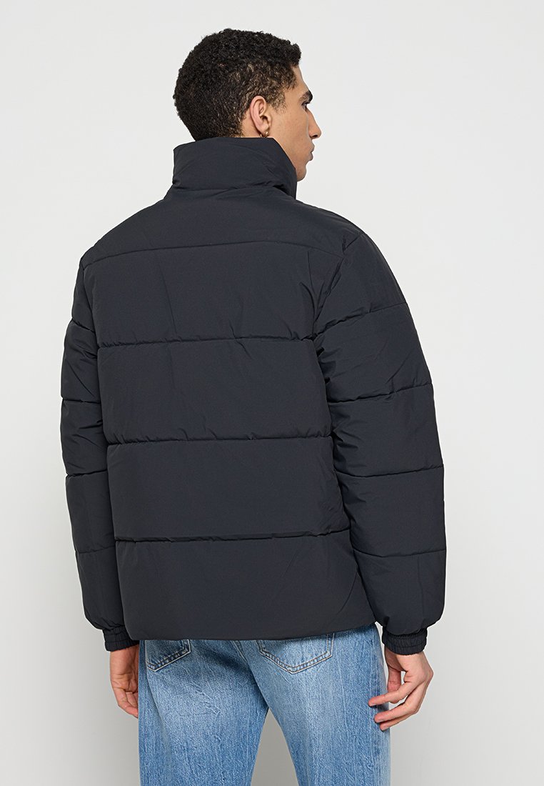 Hombre con una chaqueta de plumas negra y jeans azules, de pie con su espalda hacia la cámara frente a un fondo sencillo.