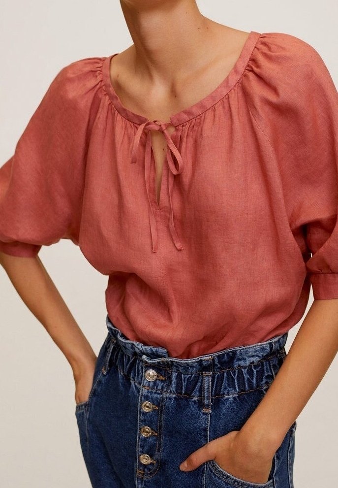 Blusa in lino rosa con scollatura rotonda, dettaglio con fiocco e maniche a sbuffo, abbinata a pantaloni denim blu a vita alta con chiusura a bottoni.