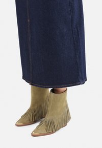 Bottines en daim vert olive avec des détails en franges et des embouts métalliques, associées à une longue jupe en denim foncé présentant des coutures visibles.