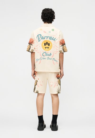 Άτομο που φοράει σετ μπεζ με κοντομάνικο πουκάμισο και σορτς με εκτυπώσεις κάκτων και το κείμενο «Barrow Club Bad Vibes, Good Times» στην πλάτη, μαύρα παπούτσια και κάλτσες.