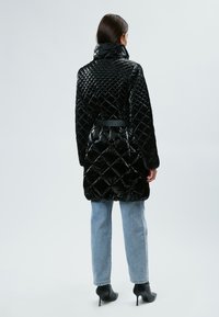 Abrigo acolchado negro con un alto collar y cintura ceñida. Presenta un material brillante y un patrón geométrico. Combinado con jeans de un color azul claro.