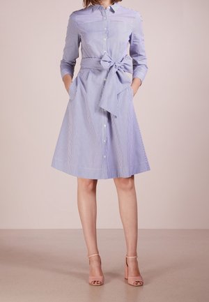 Femme portant une robe chemise à rayures verticales bleues et blanches avec un nœud à la taille et des sandales roses à talons blocs, debout les mains dans les poches.