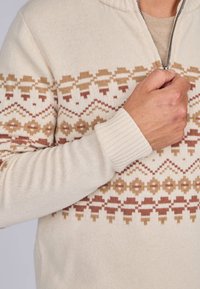 Pull en tricot couleur crème avec une texture douce. Il présente un motif géométrique dans des tons chauds de beige et de rouille. Comprend un col zippé et des poignets côtelés.