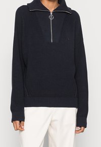 Pull en maille bleu marine avec un col zippé, un ourlet côtelé et des manches larges. Présente un corps texturé avec un design de col distinctif.