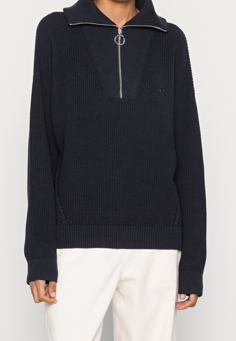 Pull en maille bleu marine avec un col zippé, un ourlet côtelé et des manches larges. Présente un corps texturé avec un design de col distinctif.