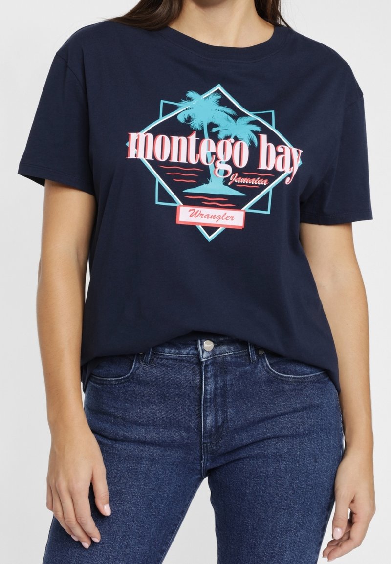 Námořnická modrá tričko s textem "Montego Bay Jamaica", grafickou palmou v tyrkysové barvě a logem "Wrangler", nošeno s tmavě modrými džínami.