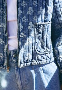 Vatteret denimjakke med blomstermønster i lyseblå, med lynlåslukning, en lomme med lap, og ribbet tekstur. Kombineret med en hvid top.