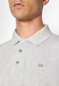 Világosszürke pólóing, texturált felülettel, gallérral, három gombbal és egy kis hímzett "CK" logóval a mellrészen.