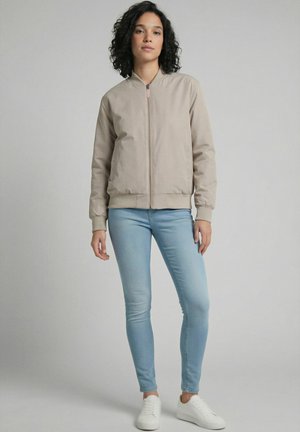 Frau mit lockigem Haar, die eine beige Jacke mit Reißverschluss, hellblaue Skinny-Jeans und weiße Sneakers trägt, steht vor einem schlichten Hintergrund.