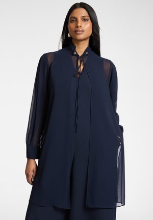 Vrouw met lang donker haar draagt een doorschijnende marineblauwe blouse met lange mouwen en een bijpassend mouwloos vest, poseert tegen een effen achtergrond.