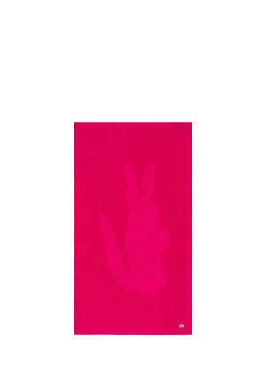 Serviette rose vif avec un grand logo crocodile ton sur ton au centre et un petit logo crocodile blanc et vert dans le coin inférieur.