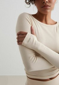 Beige långärmad topp i ribbad väv med rundad halsringning. Har tumhål vid ärmsluten och en diskret metalltagg som detalj.
