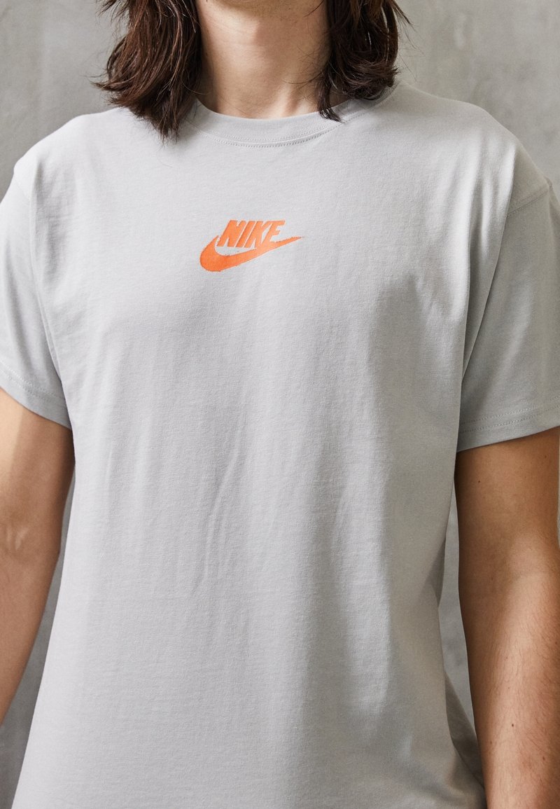 Person wearing a light gray Nike t-shirt with a bright orange logo centered on the chest, standing against a plain background.