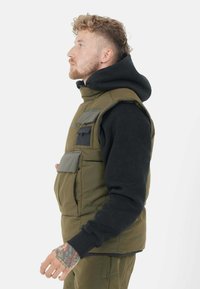 Chaleco verde oliva con un cuello alto y múltiples bolsillos, superpuesto a una sudadera con capucha negra. El chaleco tiene una textura acolchada y costuras visibles.