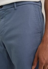 Világoskék, elegánsan szabott dressznadrág sima textúrával, lapos elejével, ferde zsebekkel és egy gombos záródással.
