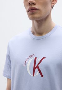 Ανοιχτό μπλε βαμβακερό μπλουζάκι με έντονο κόκκινο και λευκό λογότυπο Calvin Klein. Κοντά μανίκια και κλασικός στρογγυλός γιακάς.