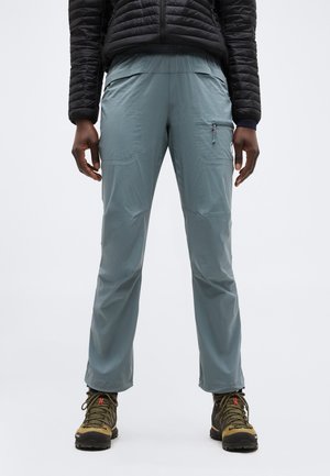 Person trägt hellblaue Wanderhose mit Reißverschlusstasche an der Seite, schwarze gesteppte Jacke und braune Wanderschuhe, steht vor weißem Hintergrund.