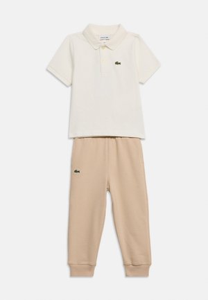 Bela polo majica s kratkimi rokavi in gumbi ter bež hlače z elastičnim manšetom, obe z majhnimi zelenimi krokodilskimi logotipi.
