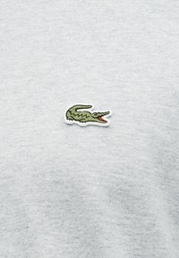 Πράσινο κεντημένο λογότυπο κροκοδείλου με ανοιχτό στόμα σε γκρι ανοιχτό ύφασμα, τοποθετημένο κεντρικά στην περιοχή του στήθους.