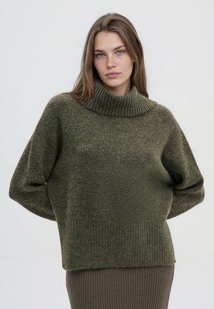 Giovane donna con capelli castani lisci che indossa un maglione a collo alto verde oliva largo e una gonna a coste, in piedi davanti a uno sfondo semplice.